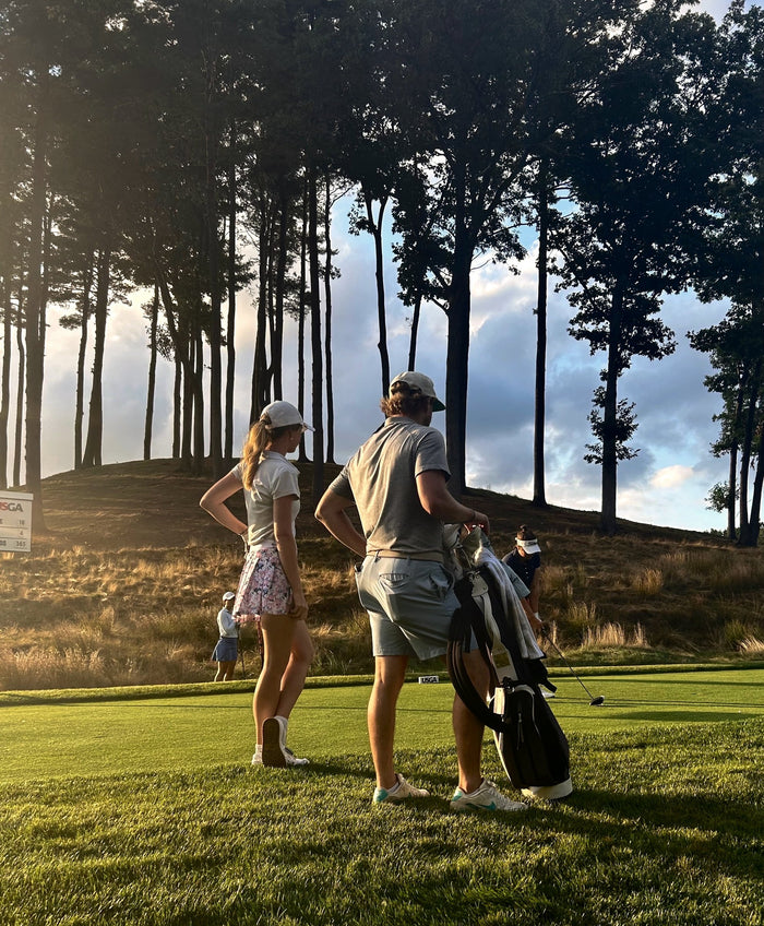 Two golfers stand together on a tee box framed by tall pines